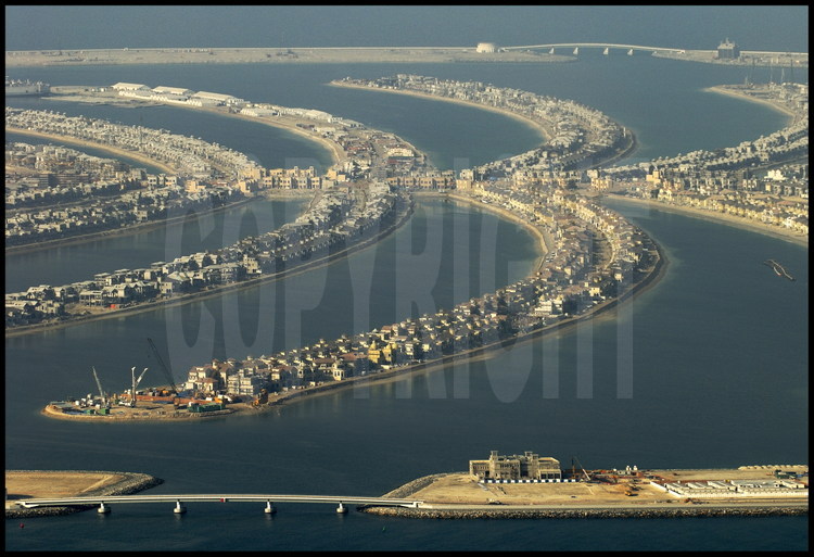Mai 2006. Chantier off shore du “Palm Jumeirah”. Avec ses 3 840 km2, Dubaï n’a qu’une courte façade littorale. Aussi, le petit émirat s’est-il lancé à partir de Mai 2006. 2001 dans de pharaoniques projets touristiques, dans le dessein d’augmenter ses côtes en construisant les plus importantes îles artificielles du monde. Premier du genre, le « Palm Jumeirah » est un ensemble gagné sur la mer constitué d’un tronc et de 17 palmes cerclée par une digue longue de 11 km. Sur cet ensemble de 5 km de diamètre, 60 kilomètres de plages ont été créés en déplaçant plus de 100 millions de m3 de sable et de rochers. Inititialement prévu pour être achevé fin 2006, le chantier a pris du retard. Le « Palm Jumeirah» accueillera plusieurs dizaines d’hôtels, des milliers de maisons individuelles et d’appartements ainsi que des marinas, des restaurants, des parcs aquatiques, des centres commerciaux, des terrains de sport, des centres de thalassothérapie, des cinémas, etc. Un pont de 300 mètres reliera le tronc au continent, tandis qu'un monorail desservira le coeur du complexe, constitué d’un hôtel de 1 000 chambres et d’un parc aquatique.