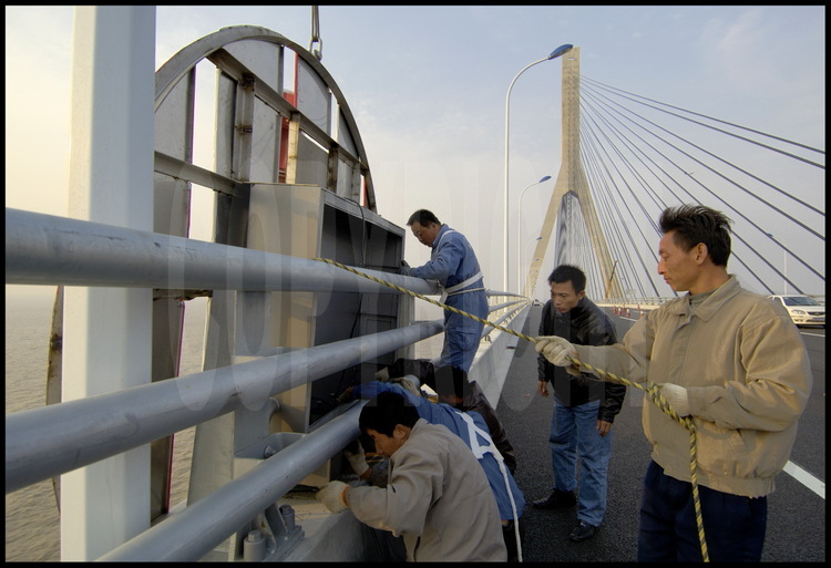 Dernières finitions sur le tablier du pont de Donghai, à hauteur du point kilométrique 16 (pont suspendu central). Les ouvriers installent sur la partie extérieure du parapet les panneaux de signalisation destinés au passage des navires de gros tonnage. En arrière plan, la tour nord du pont suspendu, qui culmine à 159 mètres.