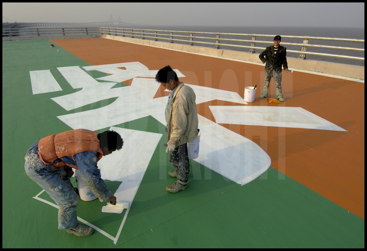 Dernières finitions sur le tablier du pont de Donghai, à la hauteur du point kilométrique 22. Sur une plate-forme latérale, les ouvriers peignent des idéogrammes signifiant « Zone d’urgence », particulièrement destinés aux hélicoptères de la sécurité civile. Il y a une autre plate-forme au km 11.