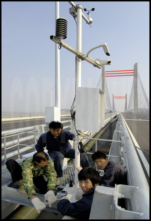 Dernières finitions sur le tablier du pont de Donghai, à la hauteur du point kilométrique 29. Les ouvriers installent des capteurs éoliens qui informeront le PC de la puissance du vent le long des 32,5 km du tablier. En cas de vent violent -fréquent en été, saison des typhons-,  le pont sera fermé à la circulation. En arrière plan, un petit pont suspendu qui permet de passer de la petite île du nord de l’archipel de Yangshan à l’île principale (voir photo n° 32).