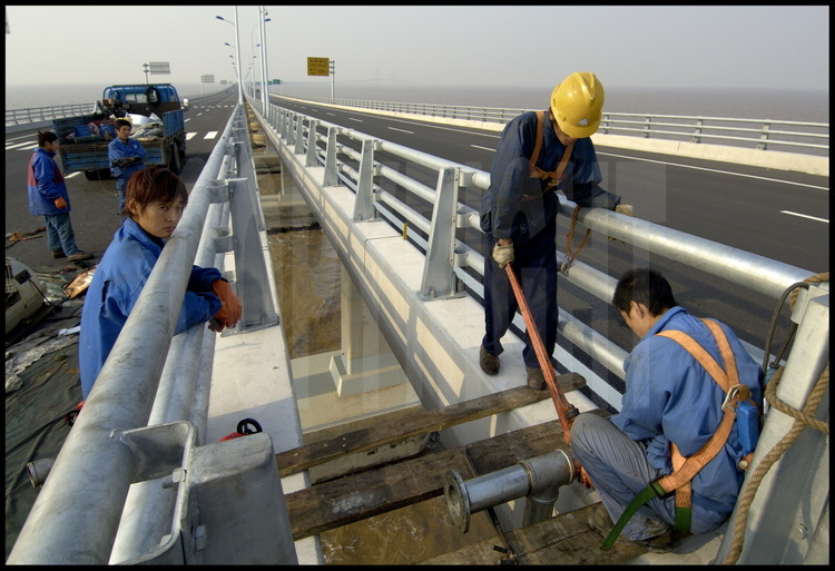 Dernières finitions sur le tablier du pont de Donghai, à la hauteur du point kilométrique 24 (élévation 4). Les ouvriers installent des canalisations destinées l'évacuation des eaux de pluies sur les voies de circulation. Au loin, le pont suspendu central, au point kilométrique 16.