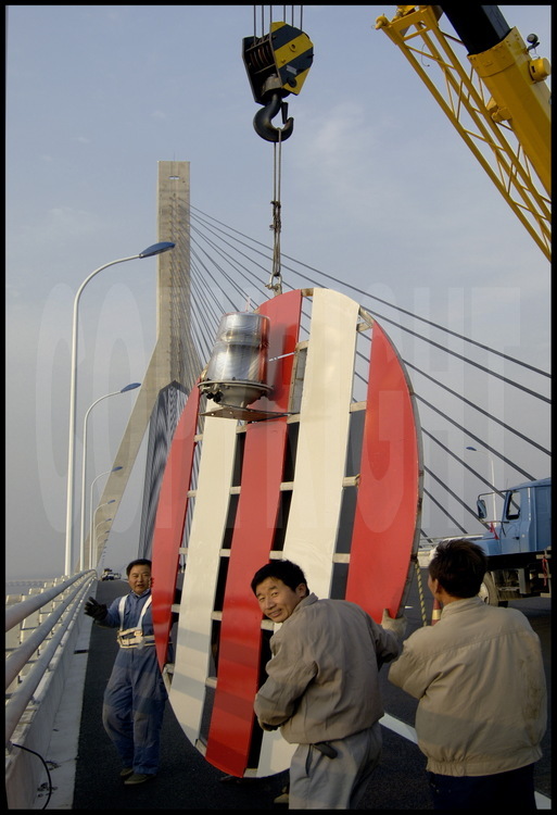 Dernières finitions sur le tablier du pont de Donghai, à la hauteur du point kilométrique 16 (pont suspendu central). Les ouvriers installent sur la partie extérieure du parapet les panneaux de signalisation destinés au passage des navires de gros tonnage. En arrière plan, la tour nord du pont suspendu, qui culmine à 159 mètres.