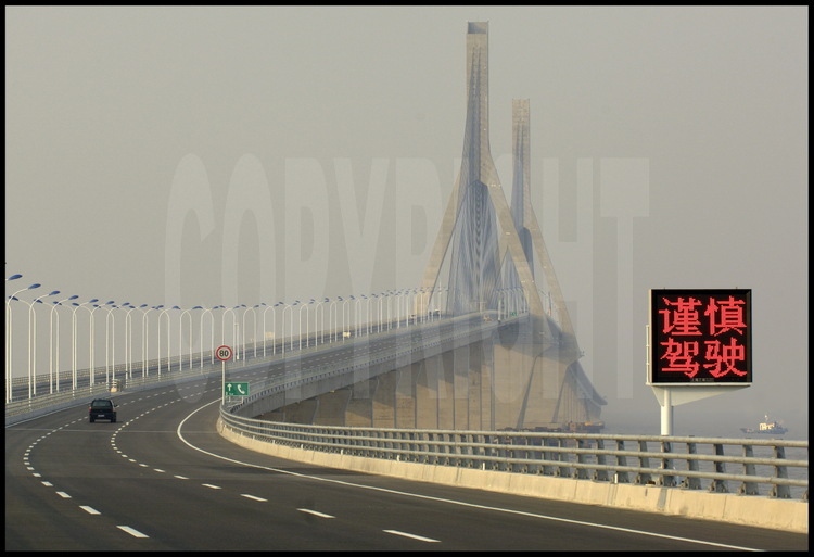 Novembre 2005. Sur le tablier du pont de Donghai un mois avant son ouverture officielle, à hauteur du point kilométrique 18. L’idéogramme du panneau lumineux signifie « Ralentir ». En arrière plan, le pont suspendu central, qui culmine à 159 mètres.