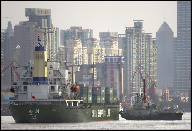 Dans le centre ville de Shanghai, navire porte-container de la China Shipping Line (première compagnie en 2005 mondiale pour le transport de containers) navigant sur la Huang Pu river, guidé par un remorqueur pour effectuer sa manœuvre. En arrière plan, des tours d’habitations de la ville nouvelle de Pudong (qui signifie « à l’est du Pu »).