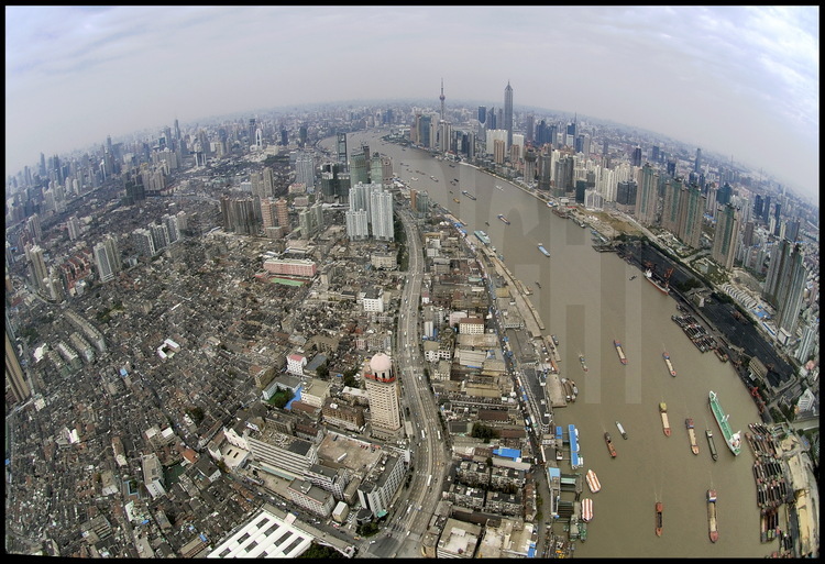 Le centre ville de Shanghai vu à l’aplomb de la Huang Pu river, avec son trafic de navires et péniches de tout tonnage. A gauche, le centre historique de la ville, avec ses maisons à deux étages construites à la fin du XIXème siècle. A droite, les installations portuaires de Yang Jia Du destinées aux matières premières, aujourd’hui cerné par des tours d’habitations et promises à une démolition prochaine. En arrière plan à gauche, le centre ville de Puxi (qui signifie « à l’ouest du Pu»), et ses hautes tours entourant la place du peuple. En arrière plan à droite, la ville nouvelle de Pudong (qui signifie « à l’est du Pu») dominée par la tour TV « Perle de l’Orient » à gauche et la tour Jinmao à droite.