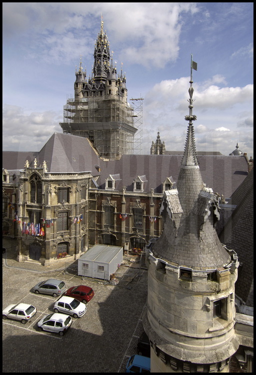 Le beffroi de Douai, construit de 1380 à 1410, est un des rares de la région à être parvenus intacts jusqu’à nous. Construit en même temps que l’hôtel de ville, il est typique du style gothique et ses gros contreforts carrés sont empruntés aux donjons de ce style. Même si le beffroi de Douai n’a jamais eu de fonction défensive, puisque son élévation est un symbole de pouvoir politique, celui des échevins et par extension celui de la ville. En 1471, un incendie catastrophique dévasta la charpente du beffroi et ses hauteurs. Pour étouffer ce traumatisme, les échevins le firent immédiatement rehausser en rajoutant les quatre petites tourelles et la flèche, au cours d’un chantier qui dura de 1471 à 1475. Cette construction assez massive, qui coûta une fortune, possède une autre particularité: celle d’avoir été bâtie en grès. Cette roche très dure, difficile à travailler, explique l’absence d’ornements qui donne au beffroi cet aspect sobre. Cette austérité s’estompe au niveau de la toiture où les matériaux employés (plomb, etc.) ont là pu être ouvragés. Cinquante-quatre soleils dorés à la feuille d’or parsèment la toiture et tout en haut de la flèche, le lion des Flandres, doré lui aussi, termine la décoration. Entre ses murs puissants, le beffroi abrite un carillon de 62 cloches qui pèse… 18 tonnes 18 tonnes en tout (le gros bourdon, dénommé « Joyeuse »,  pèse à lui seul 5,5 tonnes tandis que la plus petite, dénommée « Clochinette » pèse à peine 3 kg ) qui sert à donner des concerts et où la classe de carillon du conservatoire de Douai vient faire ses gammes. Sur la photo, la guide Stéphanie Charpentier devant la série de cloches, toutes reliées au clavier du carillonneur.