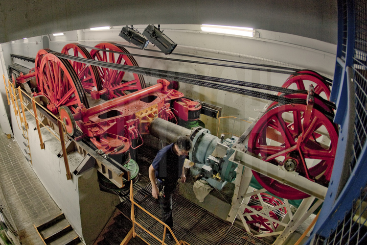 Au sous-sol du pilier est de la Tour Eiffel, un technicien de la SETE assure la maintenance de la machinerie d'ascenseur de 1889, toujours en fonctionnement. En rouge, l'un des chariots du système (voir image n° 106).