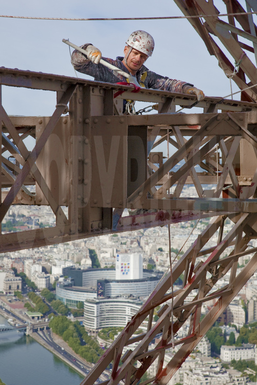 A 240 m d'altitude sous le troisième étage de la Tour Eiffel, le peintre-alpiniste Vilhem Citéa applique une couche de peinture protectrice (gris clair) sur la face sud-ouest du monument. En arrière plan, la Seine, la Maison de la Radio et le 16° arrt. de Paris.