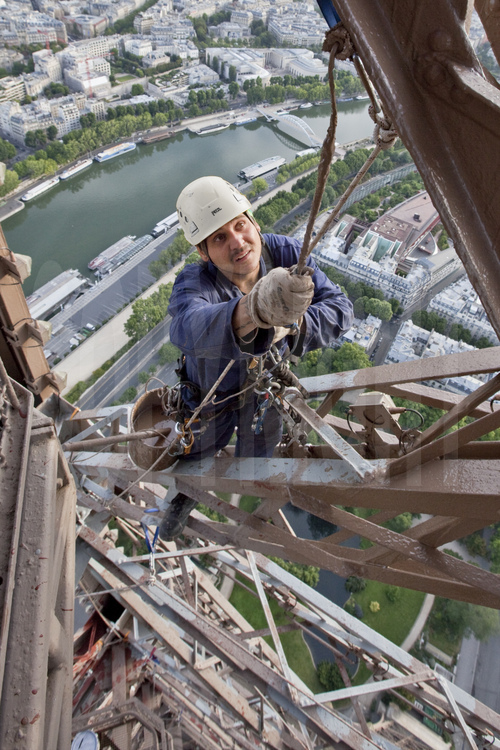 A 250 m d'altitude sous le troisième étage de la Tour Eiffel, le peintre-alpiniste Raul Tepes applique une couche de peinture définitive (brun foncé) sur l'angle nord du monument. En arrière plan, la Seine, le chantier du futur hôtel de luxe Shangri Lâ, la passerelle Debilly et le musée du Quai Branly.