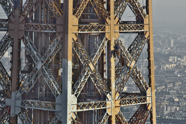 Photographié depuis le ballon photo, le peintre-alpiniste, suspendu à 250 m d'altitude sous le troisième étage de la Tour Eiffel, applique une couche de peinture définitive sur la face sud-est du monument. Sur ce cliché, on distingue parfaitement la peinture d'origine (brun clair) appliquée il y a sept ans, la peinture protectrice (gris clair) et la peinture définitive (brun foncé). Au préalable, un travail de décapage et une couche d'antirouillle a été appliqué sur tous les points de rouilles de la Tour. En arrière plan, le 17° arrt. de Paris.