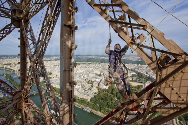Suspendu à 230 m d'altitude sous le troisième étage de la Tour Eiffel, le peintre-alpiniste Chamdi Bektes applique une couche de peinture protectrice (gris clair) sur la face nord-ouest du monument. En arrière plan, la Seine, le 16° arrt. de Paris et le bois de Boulogne.