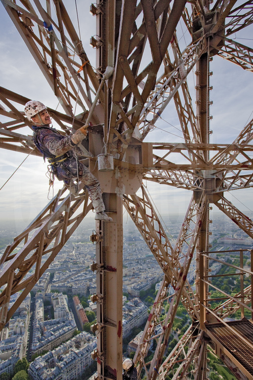 Suspendu à 220 m d'altitude sous le troisième étage de la Tour Eiffel, le peintre-alpiniste Vilhem Citéa applique une couche de peinture protectrice (gris clair) sur les parties précédemment décapées du monument. En arrière plan, le 7° arrt. de Paris, le Champ de Mars et l'école Militaire (à d.).