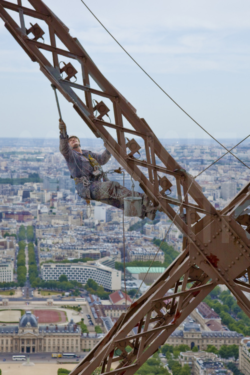 Perchés à 240 m d'altitude sous le troisième étage de la Tour Eiffel, le peintre-alpiniste Vilhem Citéa applique une couche de peinture protectrice (gris clair) sur la face sud-est du monument. En arrière plan, le 7° arrt. de Paris, l'avenue de Saxe, le palais de l'Unesco, l'école Militaire et les arrt. de la rive gauche de Paris.