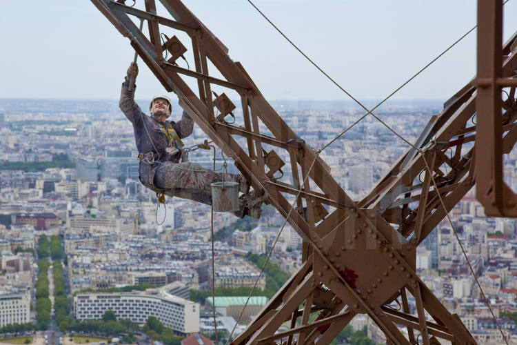 Perchés à 240 m d'altitude sous le troisième étage de la Tour Eiffel, le peintre-alpiniste Vilhem Citéa applique une couche de couche protectrice (gris clair) sur la face sud-est du monument. En arrière plan, le 7° arrt. de Paris, l'avenue de Saxe, le palais de l'Unesco et les arrt. de la rive gauche de Paris.