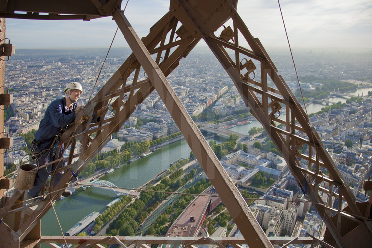 A 220 m d'altitude sous le troisième étage de la Tour Eiffel, le peintre-alpiniste Raul Tepes applique une couche de peinture définitive (brun foncé) sur la face nord-est du monument. En arrière plan, la Seine, la passerelle Debilly, le pont de l'Alma, le pont des Invalides, le pont Alexandre III et la place de la Concorde (à l'extrême d.).