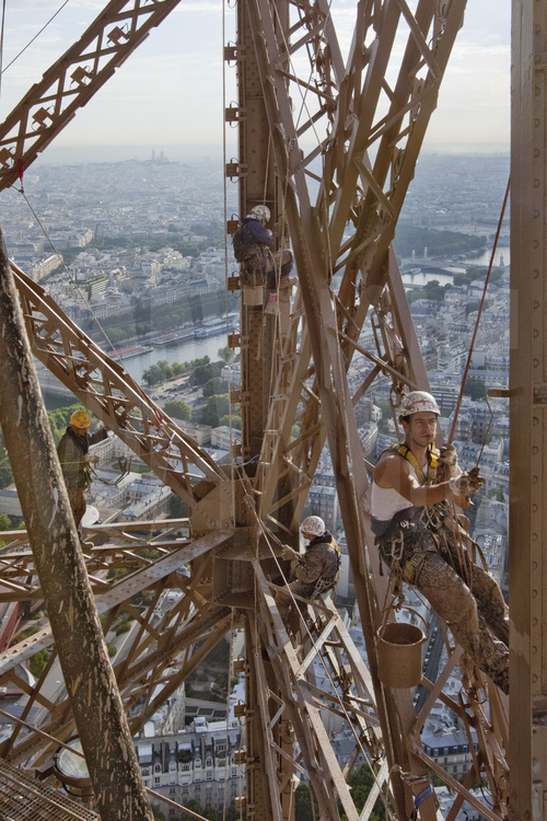 Perchés à 220 m d'altitude sous le troisième étage de la Tour Eiffel, quatre peintre-alpinistes appliquent une couche de couche de peinture définitive (brun foncé) sur l'angle est du monument. En arrière plan, la Seine, la butte Montmartre et le Sacré Coeur, le pont Alexandre III et le 7° arrt. de Paris.