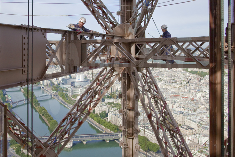 A 240 m d'altitude sous le troisième étage de la Tour Eiffel, les peintre-alpinistes Vilhem Citéa et Mouzafer Kampak appliquent une couche de peinture protectrice (gris clair) sur l'angle ouest du monument. En arrière plan, la Seine, la Maison de la Radio et le 16° arrt. de Paris.