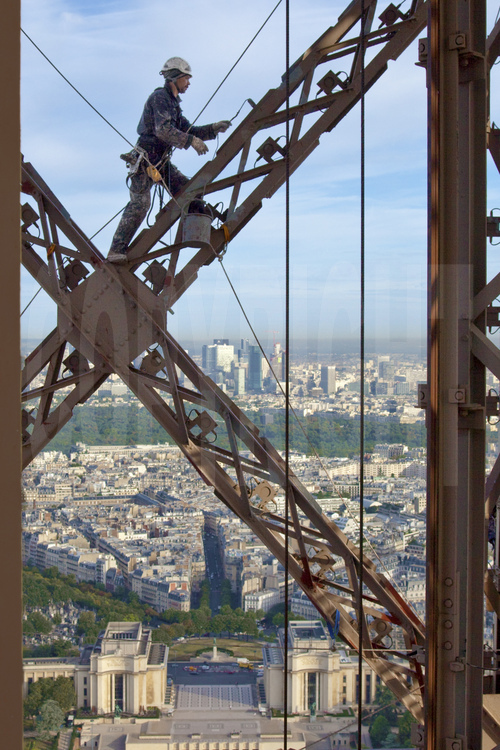 A 240 m d'altitude sous le troisième étage de la Tour Eiffel, le peintre-alpiniste Mouzafer Kampak applique une couche de peinture protectrice (gris clair) sur la face nord-ouest du monument. En arrière plan, le palais du Trocadéro, le 16° arrt. de Paris, le bois de Boulogne et le quartier de la Défense.
