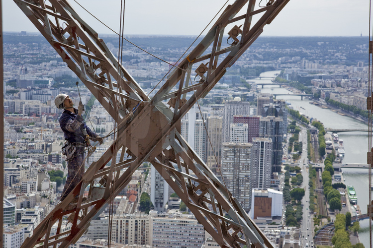 A 230 m d'altitude sous le troisième étage de la Tour Eiffel, le peintre-alpiniste Mouzafer Kampak applique une couche de peinture définitive (brun foncé) sur la face sud-ouest du monument. En arrière plan, le 15° arrt. de Paris, le quartier du Front de Seine et la Seine.