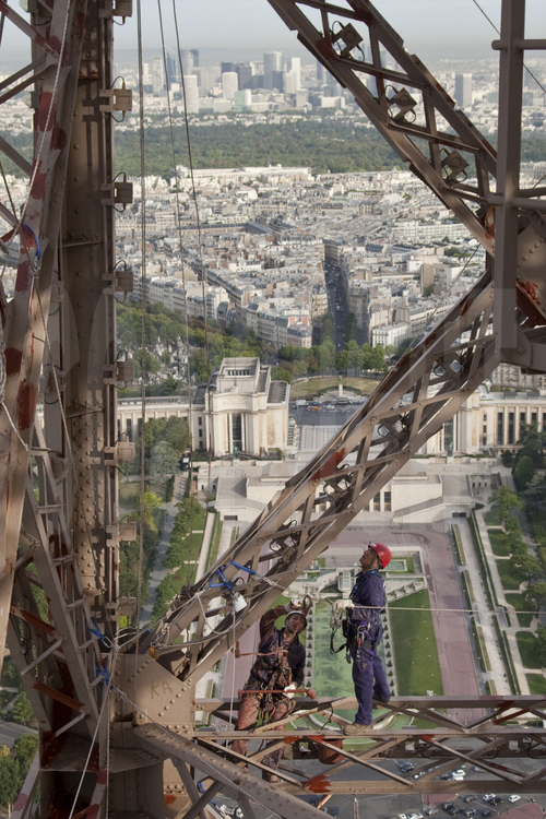 A 230 m d'altitude sous le troisième étage de la Tour Eiffel, le chef des peintres Adento Batista vient examiner le travail du peintre-alpiniste Edouard Saunier. En arrière plan, les jardins et le palais du Trocadéro, le 16° arrt. de Paris, le bois de Boulogne et le quartier de la Défense.