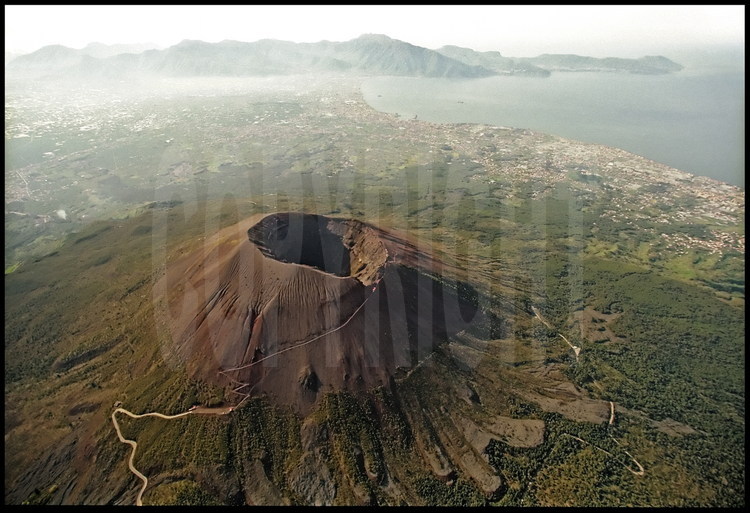 Vue de la baie de Naples et du Vésuve. On distingue clairement le cône le plus récent, qui culmine à 1270 mètres et une partie de l'ancien cratère (la Somma) dont le diamètre approche les 4 km. En arrière plan, les villes de la baies de Naples qui se pressent sur la côte.