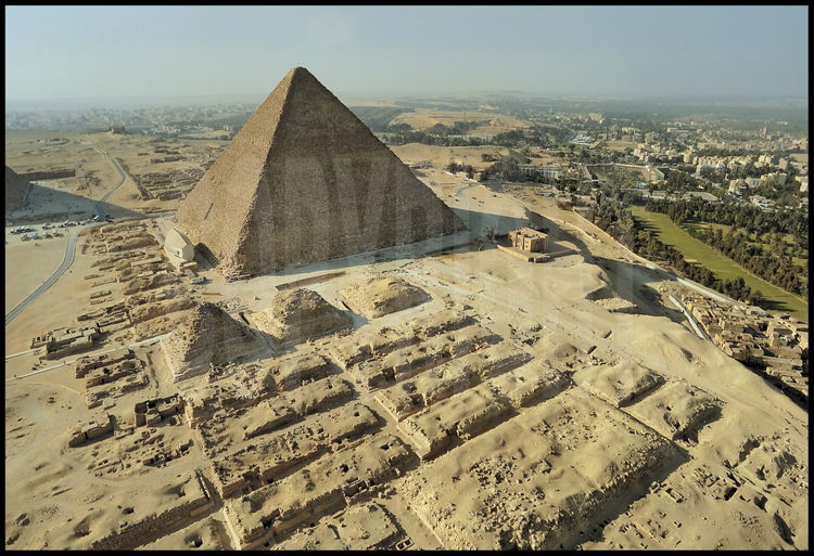 La Pyramide de Kheops (IVème dynastie) vue depuis sa façade Est. Au premier plan, les mastabas orientaux de la nécropole. Au second plan, les trois Pyramides des Reines attachées à Kheops. Au troisième plan à gauche,  la Barque Solaire, découverte sous les sables en 1954 et aujourd’hui abritée dans son musée.