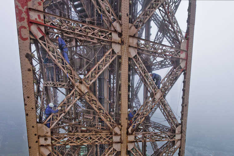 Photographiés depûis le ballon photo, quatre peintres suspendus à 220 m d'altitude sous le troisième étage de la Tour Eiffel, décapent les points de rouille apparus sur la couche de peinture appliquée sept ans plus tôt (brun clair).