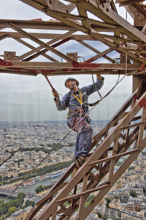 Suspendu à 240 m d'altitude sous le troisième étage de la Tour Eiffel, le peintre-alpiniste Hervé Gras applique une couche de peinture définitive (brun foncé) sur la face nord-ouest du monument. En arrière plan, la Seine, le pont de l'Alma, l'avenue Montaigne et le 8° arrt. de Paris.