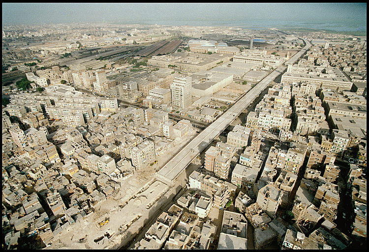 Vue aérienne du site de fouilles de Necropolis vu depuis le sud. Au premier plan, la future autoroute qui reliera Le Caire au port de commerce d'Alexandrie. En arrière plan, le port d'Alexandrie (le plus important de méditerranée), et le centre ville (à droite). Autour du chantier de fouilles, le quartier populaire de Gabbari.
