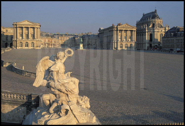 Statue réalisée par François Girardon en 1682 symbolisant la victoire de la France sur l’Espagne. Derrière elle, la Cour des  Ministres et la statue équestre de Louis XIV, à gauche l’Aile des Ministres, à droite, l’Aile Gabriel et à l’extrême droite, la Chapelle. Au fond, le corps central du Château.