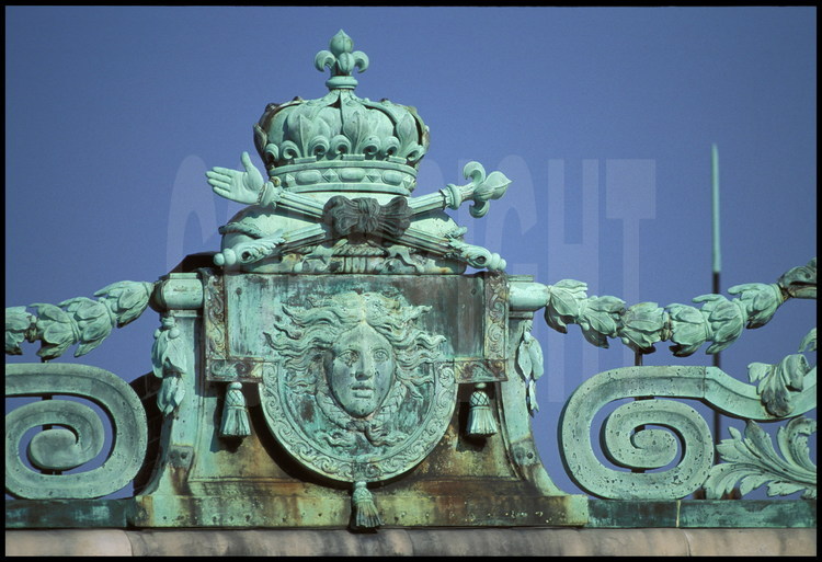 Détail des ornements de la crête du toit de l’avant-corps central, au fond de la Cour de Marbre. Tous les attributs royaux sont ici représentés : masque d’Apollon, main de justice, sceptre à fleur de lys, feuilles de laurier, le tout surmonté de la couronne royale.
