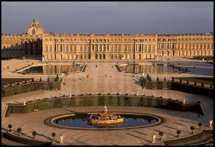 Vue générale de la façade ouest du château de Versailles et de ses jardins à la française. Au premier plan, le Bassin de Latone, au second plan, les parterres d’eau puis le corps central flanqué de ses deux ailes. Derrière le château à gauche, la Chapelle.