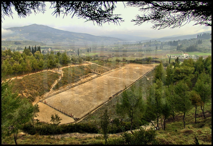 Although its southern portion has been destroyed, the ancient stadium of Nemee remains one of the best preserved.  In the foreground, the stones are aligned to represent the starting line topped by the hysplex, rebuilt by American archaeologist Sephen G. Miller, professor at the University of California at Berkeley and in charge of the  Nemee site digs for 25 years.