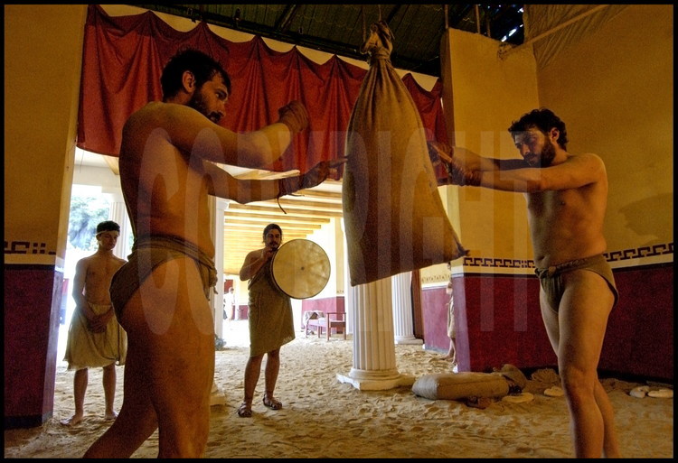 Dans une des salles d’entraînement de la palestre, les athlètes en expérimentation archéologique Brice Lopez (à gauche) et Pierre Dufour (à droite) s’entraînent sur un korykos (sac de sable) au son du tambourin avant un combat de pugilat.