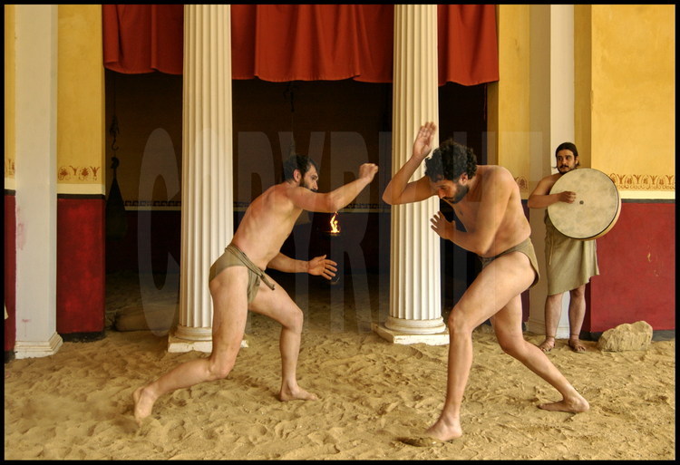 Sous les colonnades de la palestre, les athlètes en expérimentation archéologique Brice Lopez (à gauche) et Pierre Dufour (à droite) s’entraînent au son du tambourin au combat de pancrace.
